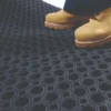A person wearing yellow work boots stands on a Roof Walkway Mat featuring a black rubber surface with a circular hole pattern.