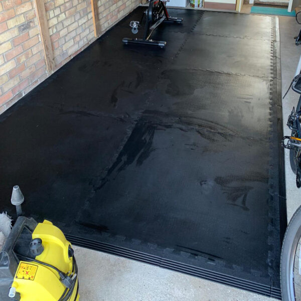 A garage space featuring black interlocking garage tile flooring, a stationary bike trainer, exposed brick wall, and a yellow vacuum cleaner on the left side.