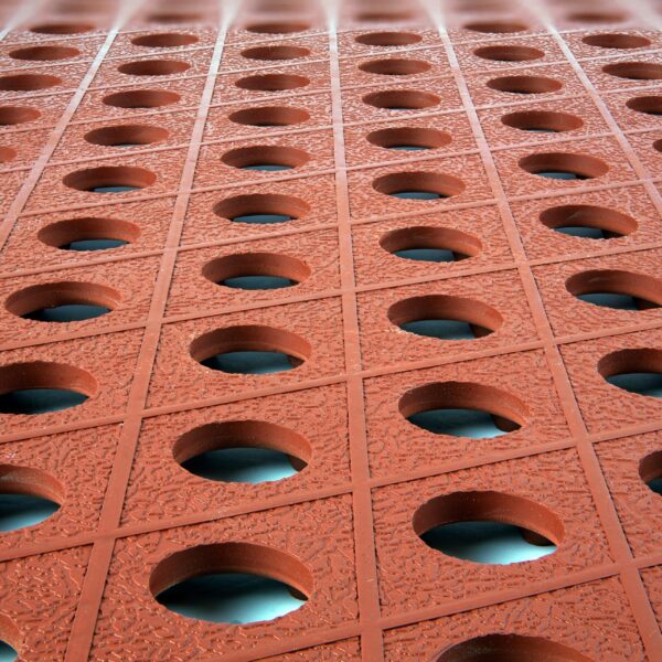 Close-up of the Food Production Mat’s textured grid of reddish-brown tiles, each featuring a central circular hole, creating a repetitive geometric pattern illuminated from above.