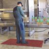 A worker in a green boiler suit and cap stands on a red Food Production Mat, packing boxes of tinned goods on an assembly line in a food processing facility.