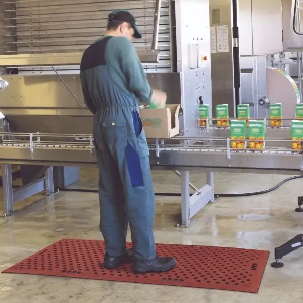 A worker in a green boiler suit and cap stands on a red Food Production Mat, packing boxes of tinned goods on an assembly line in a food processing facility.