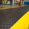 Close-up of a Circular Stud Anti-Fatigue Mat—black, textured rubber with raised circular bumps and a yellow border—on a warehouse floor. Industrial shelving and a person in a high-visibility vest appear in the background.