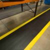 The Circular Stud Anti-Fatigue Mat, featuring a black surface and yellow border, is positioned on the floor beside a workbench or assembly line in an industrial workplace.