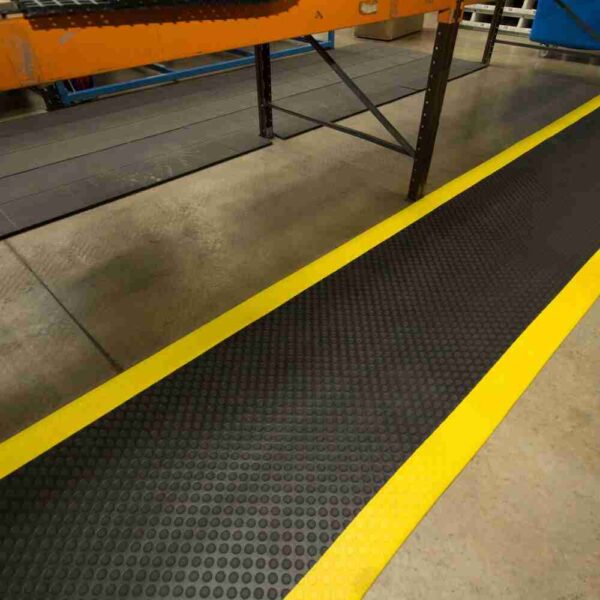 The Circular Stud Anti-Fatigue Mat, featuring a black surface and yellow border, is positioned on the floor beside a workbench or assembly line in an industrial workplace.