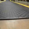 Close-up of the Circular Stud Anti-Fatigue Mat, featuring a black rubber surface with raised circular studs and yellow borders, placed on a smooth indoor concrete floor.