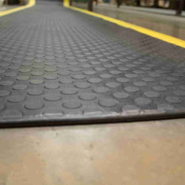 Close-up of the Circular Stud Anti-Fatigue Mat, featuring a black rubber surface with raised circular studs and yellow borders, placed on a smooth indoor concrete floor.