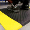 A person wearing black and grey shoes stands on a Circular Stud Anti-Fatigue Mat with bright yellow safety edges on a grey floor.
