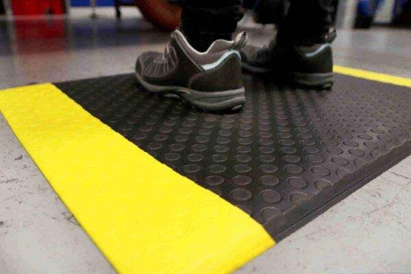 A person wearing black and grey shoes stands on a Circular Stud Anti-Fatigue Mat with bright yellow safety edges on a grey floor.