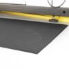 A close-up of the Ribbed Anti-Fatigue Mat in black on a white floor, placed before white cupboards with a yellow strip and black metal support leg visible in the background.