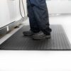 A person in black shoes and navy trousers stands on a Ribbed Anti-Fatigue Mat with a textured black surface in a clean, bright indoor space featuring light-coloured flooring.
