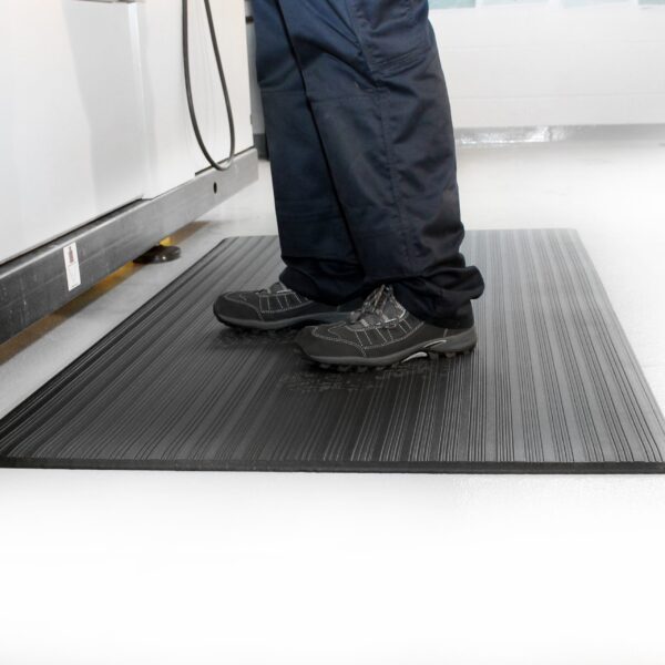 A person in black shoes and navy trousers stands on a Ribbed Anti-Fatigue Mat with a textured black surface in a clean, bright indoor space featuring light-coloured flooring.
