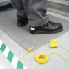 A person in black shoes and grey trousers stands on the Anti-Static Mat. Nearby, a yellow grounding lead with a clip is coiled on the floor beside green and white striped floor markings.