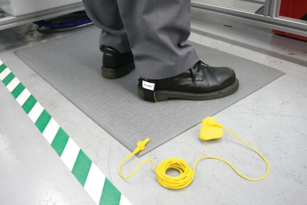 A person in black shoes and grey trousers stands on the Anti-Static Mat. Nearby, a yellow grounding lead with a clip is coiled on the floor beside green and white striped floor markings.