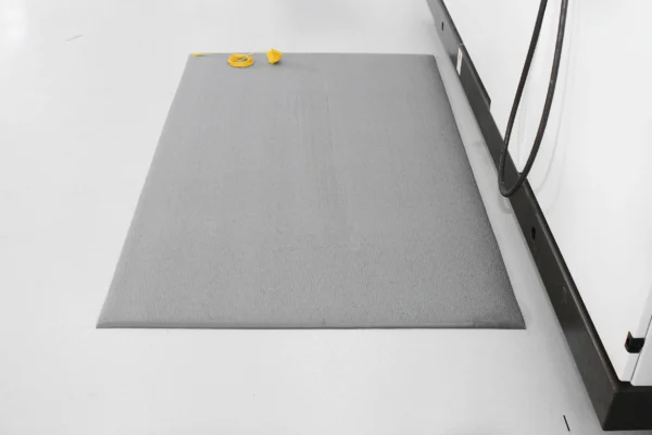 An Anti-Static Mat in grey is placed on a white floor next to a white wall with a black skirting board. A yellow measuring tape is coiled and resting near the top left corner of the mat.
