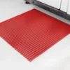 A Heavy-Duty Cross-Ribbed Mat in red rubber with a square grid pattern is placed on a light grey floor near a cupboard.