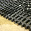 Close-up of the Heavy-Duty Cross-Ribbed Mat in black plastic on a wooden surface, showing small water droplets beneath and some debris scattered on top.