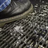 A black leather shoe steps on a Heavy-Duty Cross-Ribbed Mat, which is covered with scattered metal shavings, some of which have fallen through the mat’s openings.