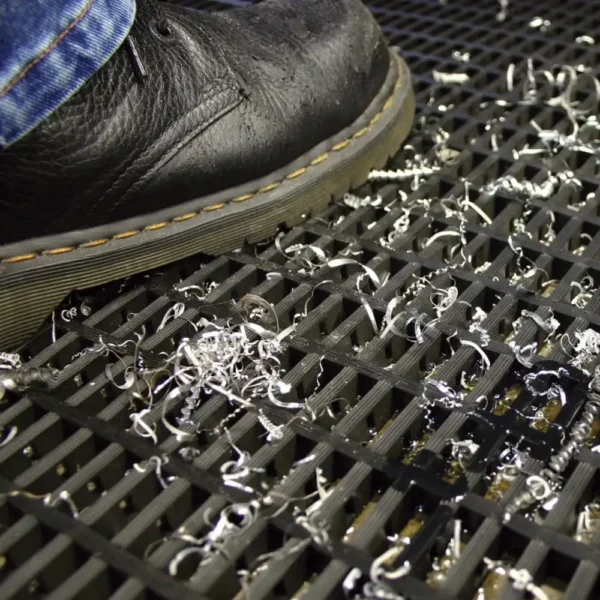 A black leather shoe steps on a Heavy-Duty Cross-Ribbed Mat, which is covered with scattered metal shavings, some of which have fallen through the mat’s openings.