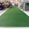 A clean, well-maintained Heavy-Duty Cross-Ribbed Mat forms a long green grated walkway through an industrial workspace lined with machinery, shelves, and workstations.