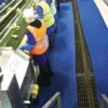 Workers in safety vests and hats sort items on a conveyor belt in a plant; the floor beside a metal grating is covered with Heavy-Duty Cross-Ribbed Matting.