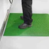 A person in black trousers and dark shoes stands on a Standard Cross-Ribbed Mat, featuring a bright green, textured anti-slip surface, placed on a light-coloured floor near a wall or appliance.