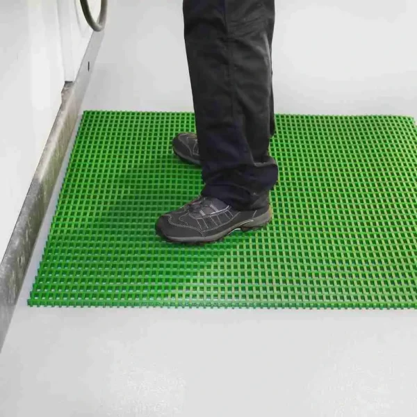 A person in black trousers and dark shoes stands on a Standard Cross-Ribbed Mat, featuring a bright green, textured anti-slip surface, placed on a light-coloured floor near a wall or appliance.
