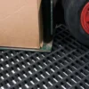 A cardboard box sits on a metal trolley with a red wheel, positioned on the black Premium Cross-Ribbed Mat.