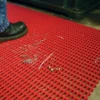 A person wearing black shoes stands on a Premium Cross-Ribbed Mat with a grid pattern, placed on an industrial floor. Metal swarf and liquid are scattered on the red anti-fatigue mat.