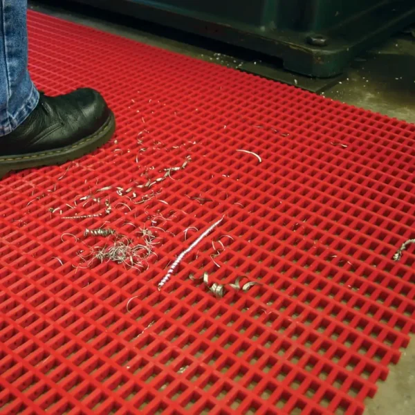 A person wearing black shoes stands on a Premium Cross-Ribbed Mat with a grid pattern, placed on an industrial floor. Metal swarf and liquid are scattered on the red anti-fatigue mat.