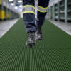 Close-up of a person wearing black work boots and navy trousers with reflective stripes, walking on Open Top Grating in an industrial setting. Shelving and bright lighting are visible in the background.