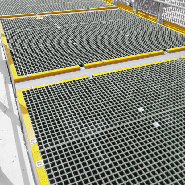 Open Top Grating with yellow safety edges is installed on an elevated platform, providing drainage and ventilation through its grid pattern; handrails in the background offer extra safety.