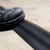 A person in black boots steps on a flexible black PVC Cable Protector (HD) placed on a concrete floor.