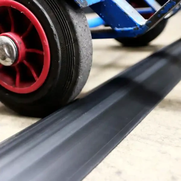A close-up of a blue trolley's red and black wheel rolling over a PVC Cable Protector (HD) on a light-coloured surface, with part of the trolley frame visible.