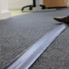 A close-up of a grey carpeted floor with a PVC Cable Protector (GP) in black running across it. Part of a brown shoe and some office furniture are visible in the background.