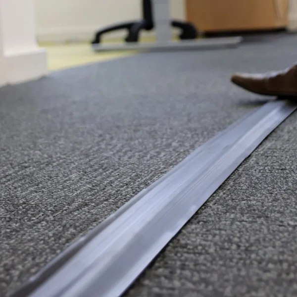 A close-up of a grey carpeted floor with a PVC Cable Protector (GP) in black running across it. Part of a brown shoe and some office furniture are visible in the background.