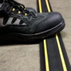 A black hiking boot with yellow accents steps on a PVC Cable Protector (GP) featuring yellow and black stripes, placed on a concrete surface.