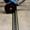 A blue sack truck wheel rolls over a black and yellow PVC Cable Protector (GP) placed on a concrete surface.