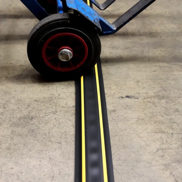 A blue sack truck wheel rolls over a black and yellow PVC Cable Protector (GP) placed on a concrete surface.