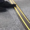 A person wearing black shoes steps near a PVC Cable Protector (GP) in black and yellow on a grey carpeted surface.