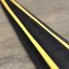 A close-up of the PVC Cable Protector (GP) in black, featuring two bright yellow stripes along its length, placed on a light brown floor.