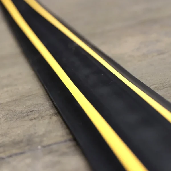 A close-up of the PVC Cable Protector (GP) in black, featuring two bright yellow stripes along its length, placed on a light brown floor.