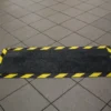The Cable Protector Mat is a rectangular black floor mat with yellow and black caution stripes around the edges, placed on a tiled floor.