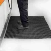 A person in dark trousers and black work shoes stands on a Standard Cross-Ribbed Mat with a grid pattern beside industrial equipment in a clean indoor setting.
