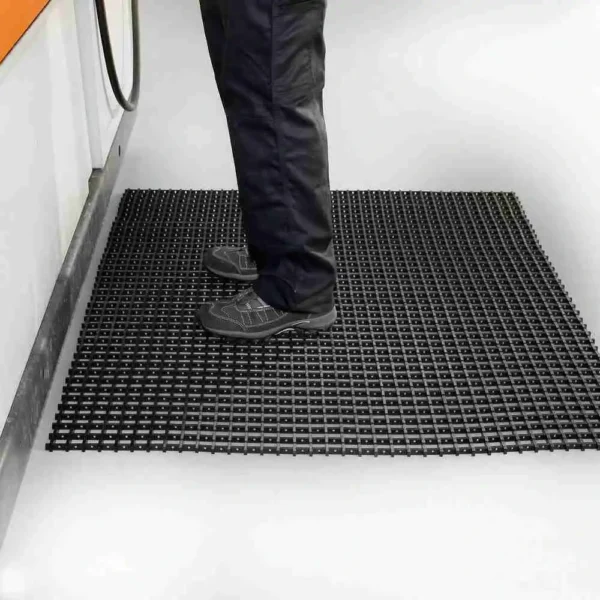 A person in dark trousers and black work shoes stands on a Standard Cross-Ribbed Mat with a grid pattern beside industrial equipment in a clean indoor setting.