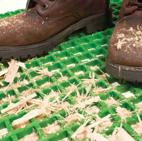 A pair of brown work boots covered in wood shavings stands on a Standard Cross-Ribbed Mat, with scattered wood shavings visible on the green mat’s surface.
