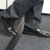 A person steps over a slightly raised PVC Cable Protector, transitioning from grey carpet flooring, while wearing black dress shoes and dark trousers.