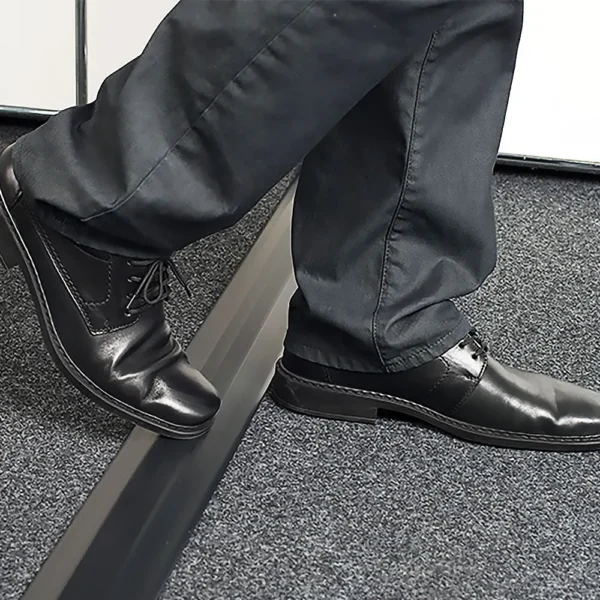 A person steps over a slightly raised PVC Cable Protector, transitioning from grey carpet flooring, while wearing black dress shoes and dark trousers.