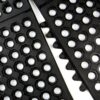 Close-up of two Interlocking Anti-Fatigue Mats in black rubber, featuring hexagonal holes and interlocking edges, arranged diagonally on a white background.