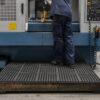 A worker in blue uniform stands on an Interlocking Anti-Fatigue Mat in front of an industrial machine, operating controls and holding a yellow handheld device. The machine has buttons, a display panel, and safety signs.