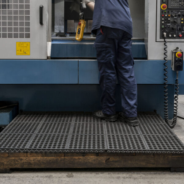 A worker in blue uniform stands on an Interlocking Anti-Fatigue Mat in front of an industrial machine, operating controls and holding a yellow handheld device. The machine has buttons, a display panel, and safety signs.
